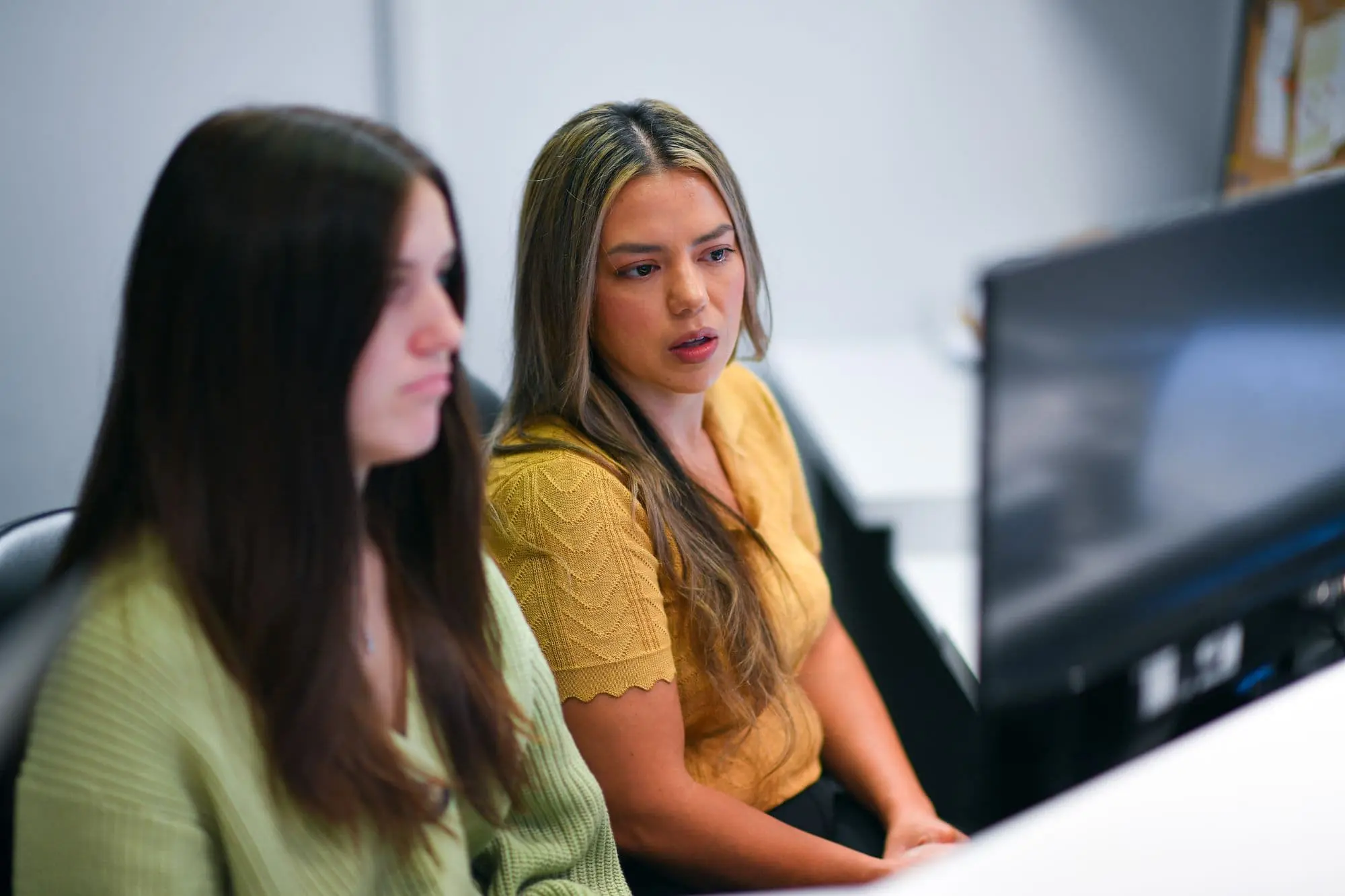 Two women, focused on a computer screen at their desk, discuss orthodontic insurance plans. One wears a green top, the other yellow, as they diligently review options with precision and care in Sammamish or Kent, WA