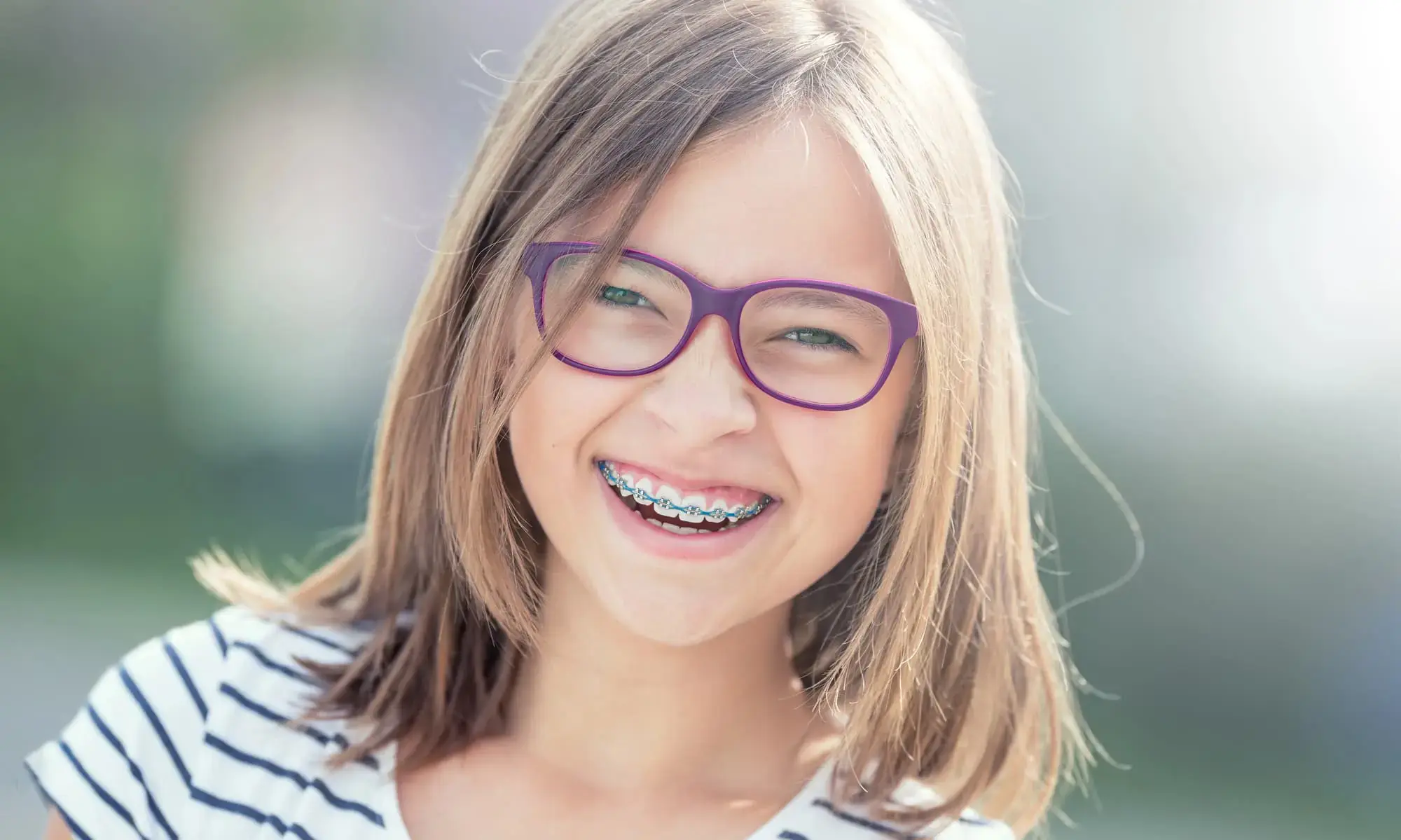 A smiling girl with glasses and metal braces, wearing a striped shirt, stands outdoors at Freelove Orthodontics, orthodontist in Sammamish and Kent, WA