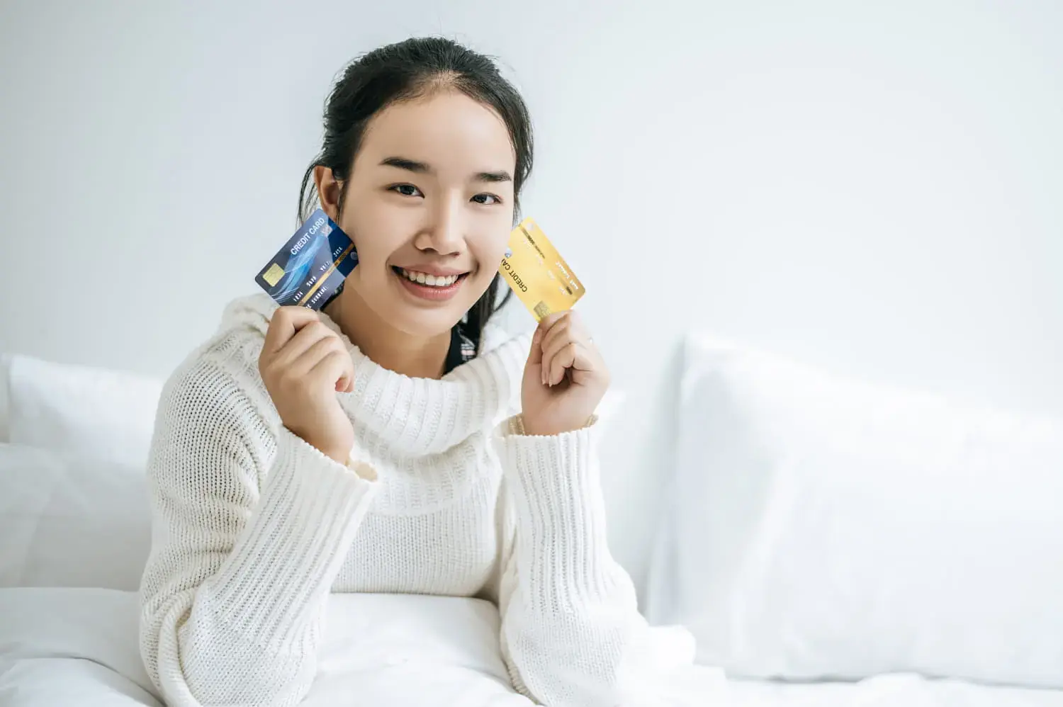 Person in a white sweater sitting on a bed, smiling while holding a blue and a yellow credit card for braces payment plan at Freelove orthodontics in Sammamish or Kent, WA