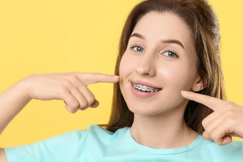 A smiling person with metal braces in Kent, WA, showcases their teeth while wearing a light blue shirt against a vibrant yellow background. Their long brown hair adds to the cheerful scene.
