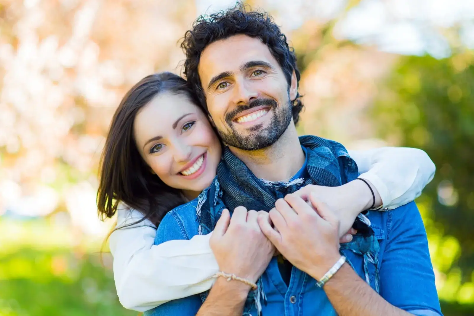 A couple, smiling and embracing in a sunlit park, showcases their happiness with gleaming smiles. The woman hugs the man from behind, both wearing casual attire. Their confidence shines through, thanks to comprehensive braces orthodontic insurance care in Sammamish or Kent WA