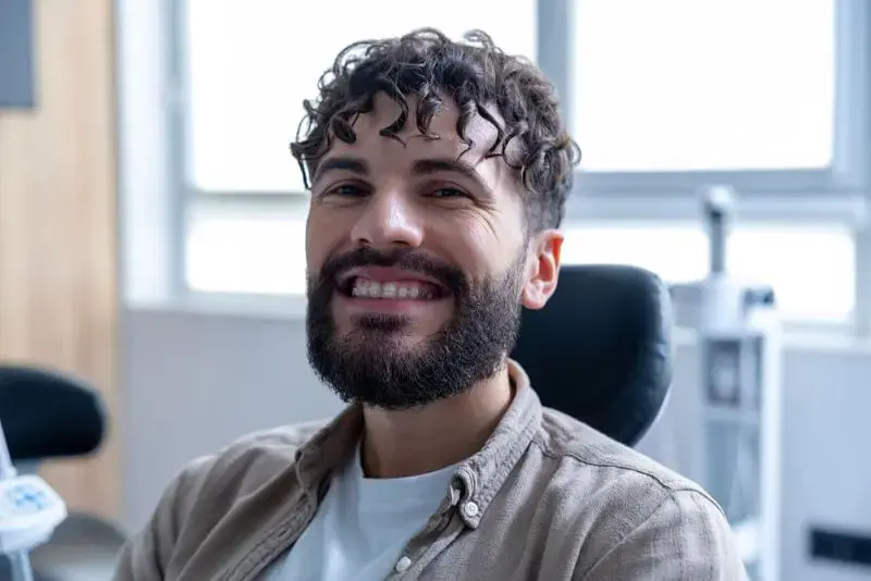 A man with curly hair and a beard smiling while sitting in a chair, wearing braces on his teeth. Background features a window and blurred office equipment. Illustrating clear braces Sammamish or Kent, WA