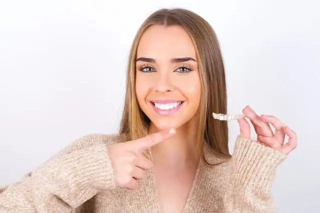 A smiling woman with long hair holds a Invisalign clear aligner in one hand and points to it with the other, showcasing her choice for braces in Sammamish, WA. She is wearing a beige sweater.