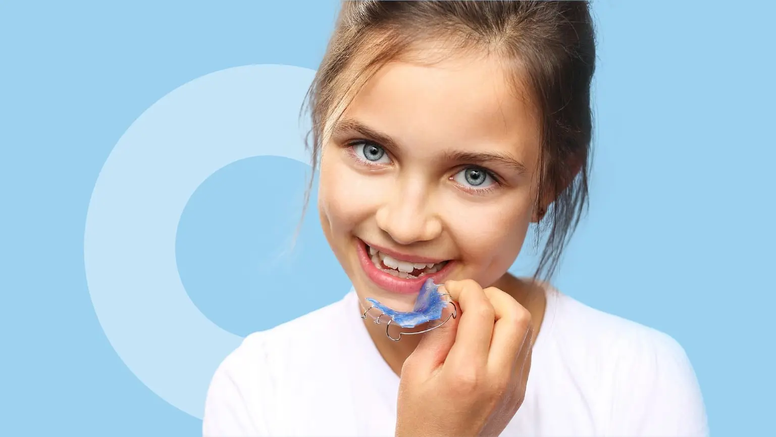 A young girl, smiling against a light blue background, holds an orthodontic appliances from orthodontist in Sammamish or Kent, WA—a blue dental retainer—near her teeth.