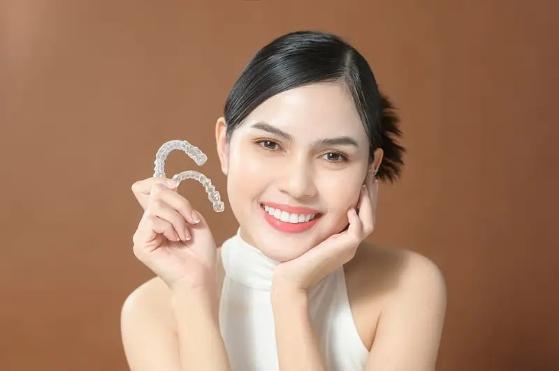 Person smiling and holding Invisalign clear dental aligners against a brown background, showcasing an alternative to traditional braces in Kent, WA.