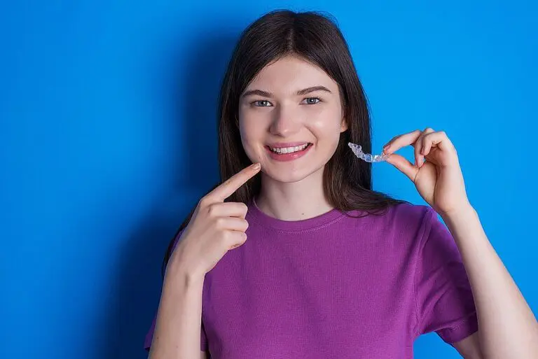 Person in a purple shirt holding a dental aligner, pointing at their smile against a blue background, showcasing one of the popular alternatives to braces in Kent and Sammamish, WA.
