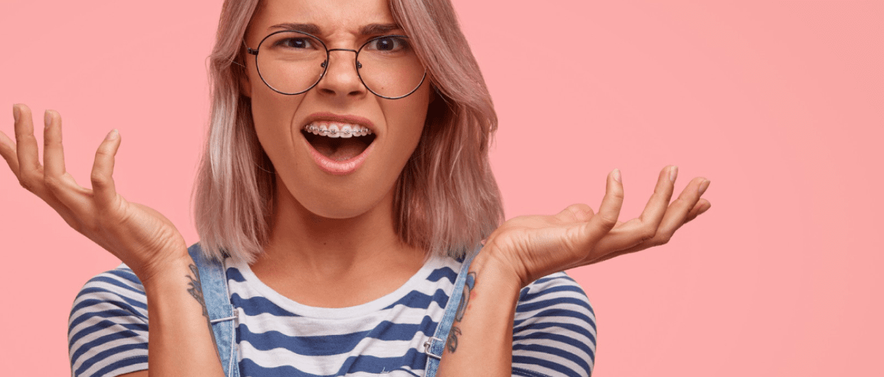 A person with glasses and adult braces, wearing a striped shirt, stands against a pink background with an open-mouthed, confused or frustrated expression and raised hands in Kent or Sammamish, WA.