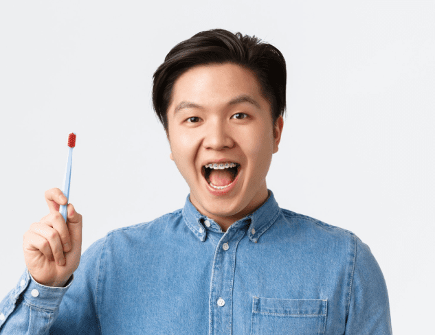 A person wearing a denim shirt smiles with an open mouth and holds up a blue toothbrush with red bristles against a plain light background in Kent or Sammamish, WA.
