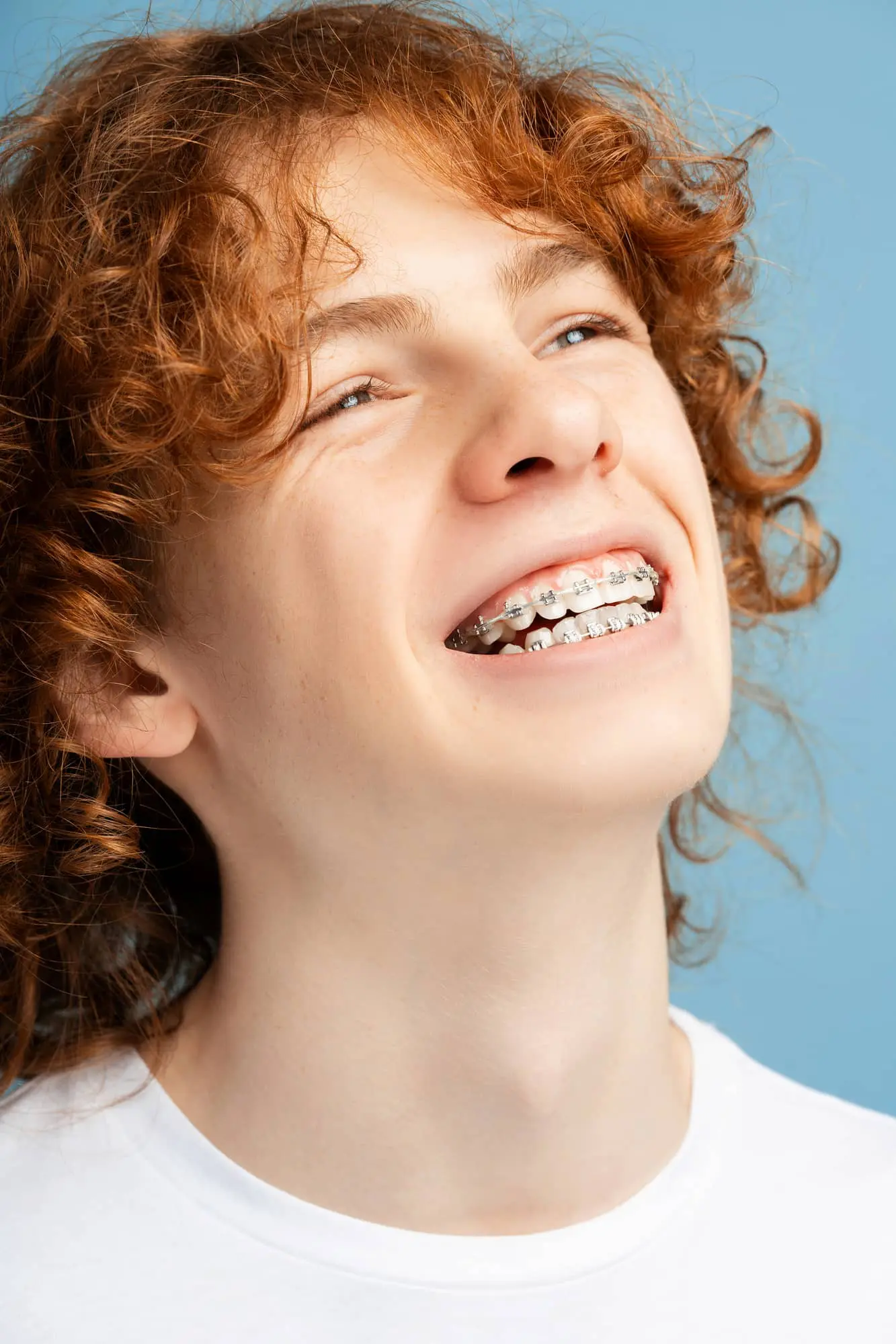 Teen with curly red hair and adult braces smiles, looking upward, against a plain blue background in Kent or Sammamish, WA.