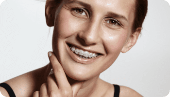 A person with light skin and brown hair smiles, showing clear braces on their teeth, and touches their chin with one finger in Kent or Sammamish, WA.
