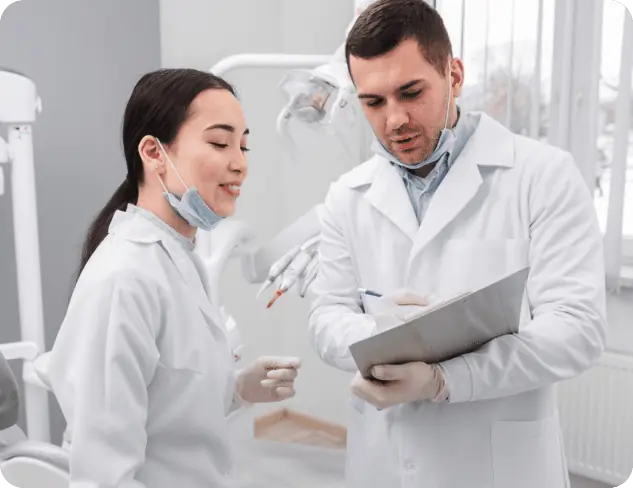 Two dental professionals in white lab coats discuss notes on a clipboard in a dental office, with dental equipment and models of adult braces visible in the background.