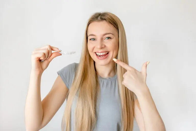 Woman with long blonde hair smiling and holding a clear dental aligner, pointing to her teeth, standing against a plain white background. Wondering: Does insurance cover Invisalign in Kent and Sammamish, WA?.