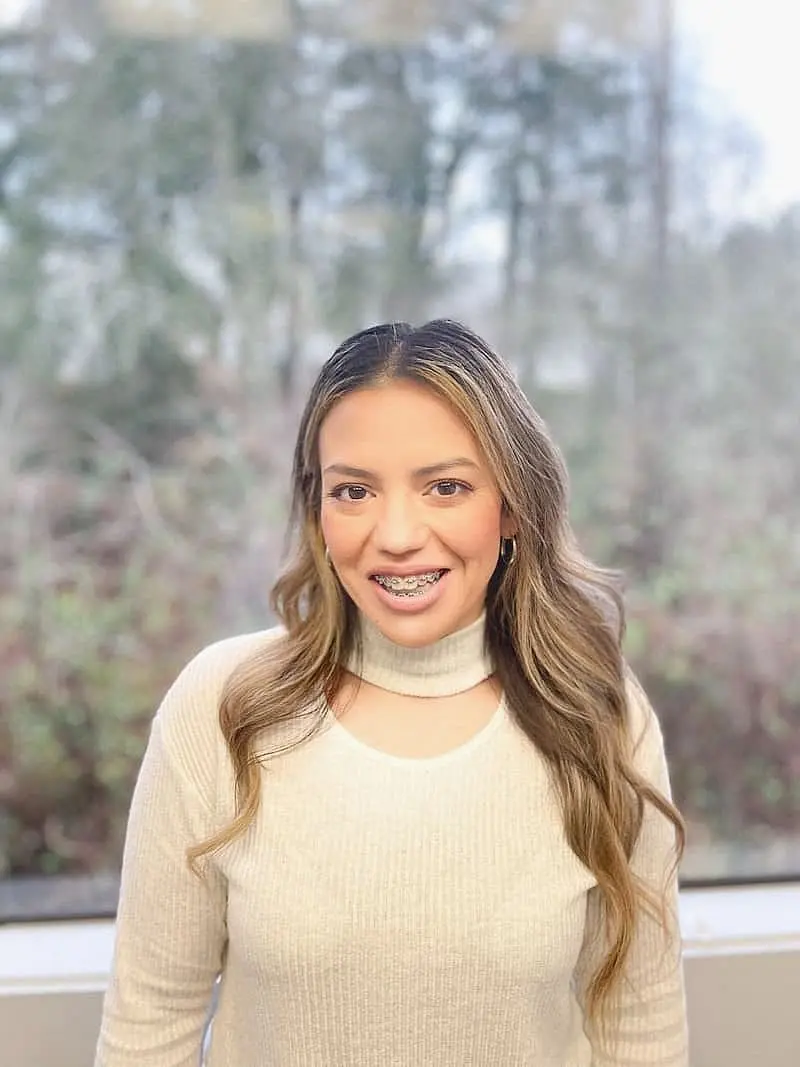 A person wearing a white top and braces from Freelove Orthodontics in Kent, WA, stands smiling in front of a window with a blurred forested background.
