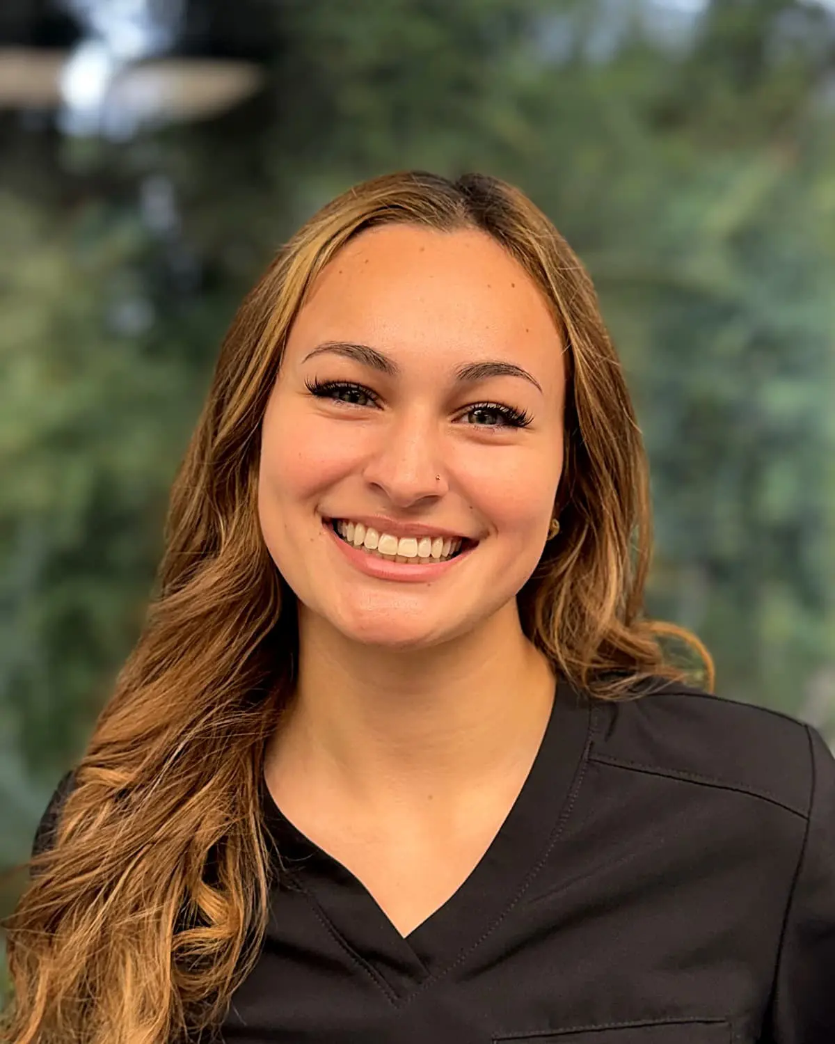At Freelove Orthodontics in Kent or Sammamish, WA, Carmen, an Orthodontic Assistant with long light brown hair in a dark scrub top, smiles in front of a blurred green outdoor background.