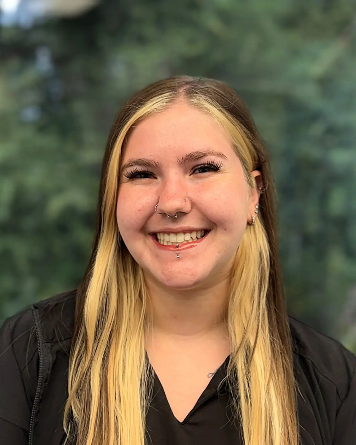 Delainey, an orthodontic assistant at Freelove Orthodontics in Kent or Sammamish, WA, smiles warmly at the camera. She has long blonde hair and facial piercings, standing outdoors with green trees softly blurred behind her.