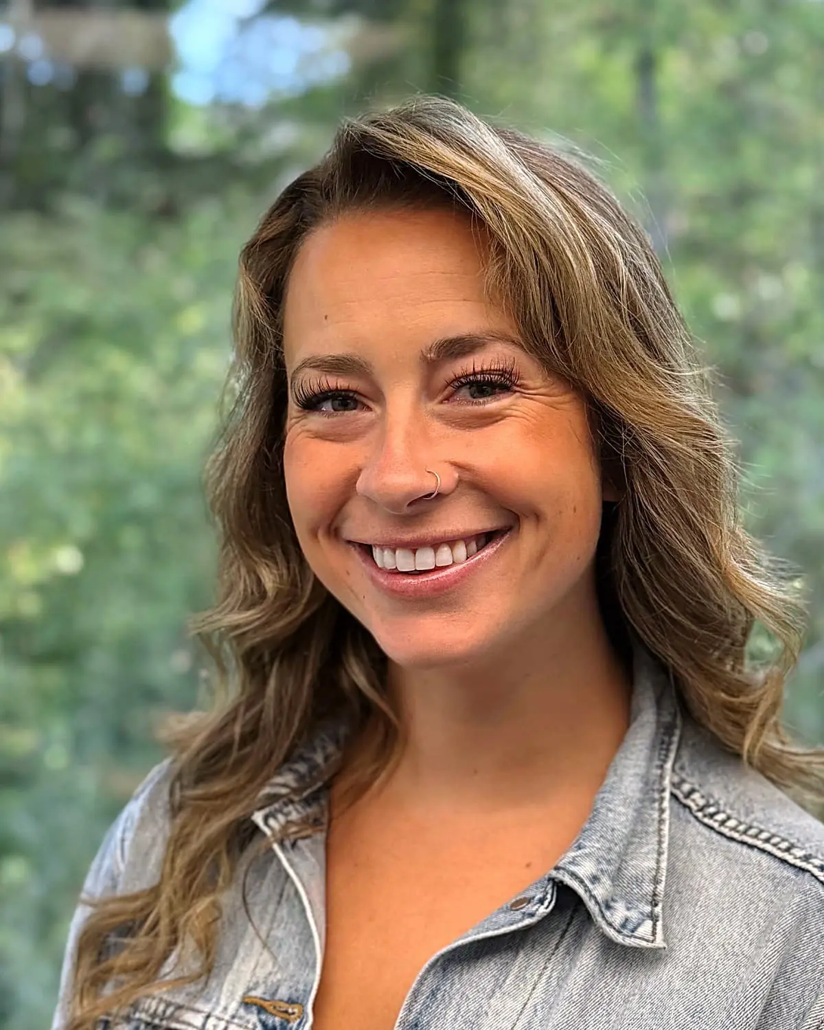 Katie, the Treatment Coordinator at Freelove Orthodontics in Kent or Sammamish, WA, smiles in a denim jacket with wavy light brown hair against a blurred green outdoor background.