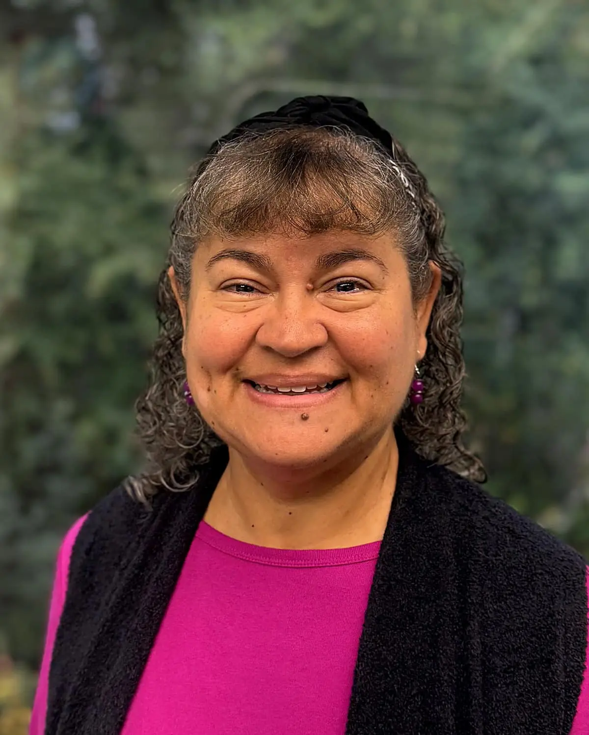 At Freelove Orthodontics in Kent or Sammamish, WA, Nicole, the Scheduling & Financial Coordinator with curly hair and a black headband, smiles at the camera in a magenta top and black vest with a blurred green outdoor background.
