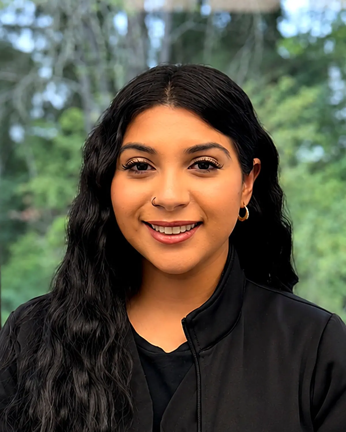 Silvia, Orthodontic Assistant at Freelove Orthodontics in Kent or Sammamish, WA, smiles at the camera with long dark wavy hair, a black jacket, and hoop earrings against a blurred green outdoor background.
