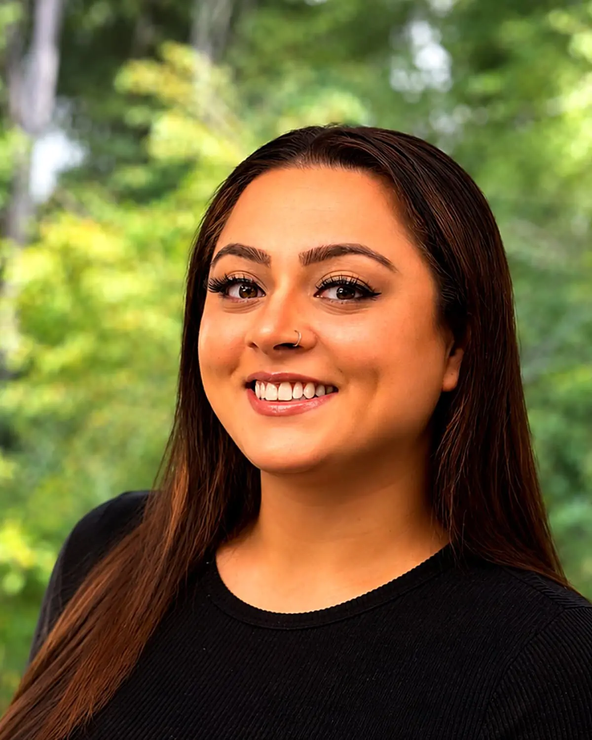 Smiling in front of a blurred green background, Victoria, Clinical Coordinator at Freelove Orthodontics in Kent or Sammamish, WA, wears a black top and has long brown hair and a nose ring.